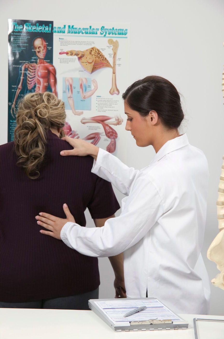 A chiropractic healthcare professional examines a patient's back in a Manhattan clinic with anatomical posters in the background. She is wearing a white coat, and the patient is in casual attire. A chiropractic healthcare professional examines a patient's back in a Manhattan clinic with anatomical posters in the background. She is wearing a white coat, and the patient is in casual attire.